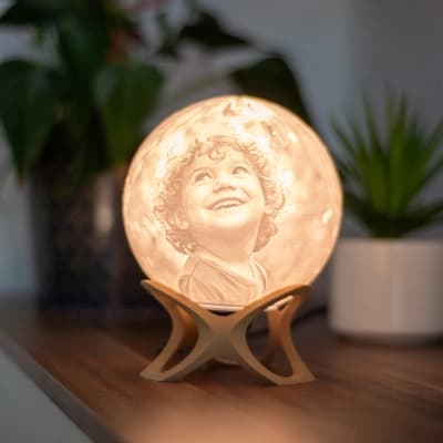 Luna personalizirana 3D foto lampa s dječjim portretom koja svijetli na drvenom postolju, postavljena na polici s biljkama.